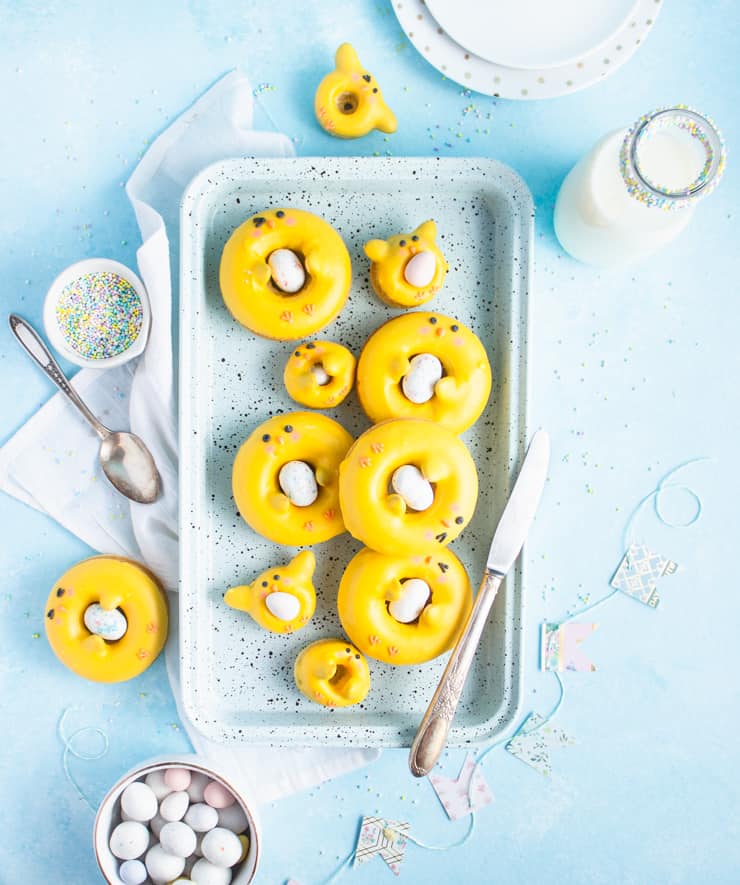 Tray of yellow donut decorated to look like chicks. Utensils, glass of milk and egg candies are strewn around the outside of the tray.